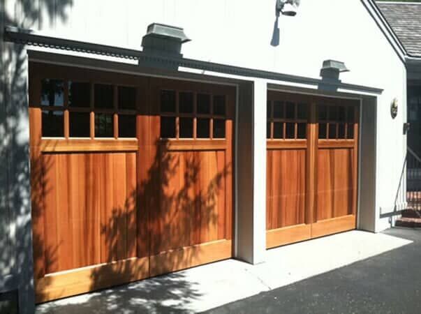 Two brown wooden garage doors with windows, mounted on a white exterior wall.