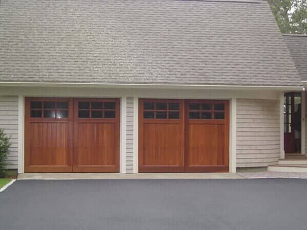 Two side-by-side wooden garage doors with windows on the top panels, attached to a suburban home with grey siding.