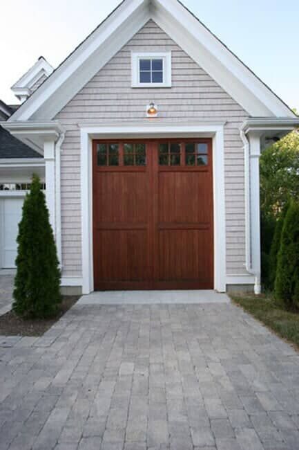 A single-car wooden garage door with windows, set in a light-colored, shingled home with a stone paver driveway.