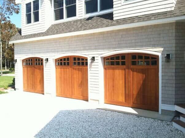 A three-car garage with arched, wood-stained carriage-style doors and windows, set in a light shingled exterior home.