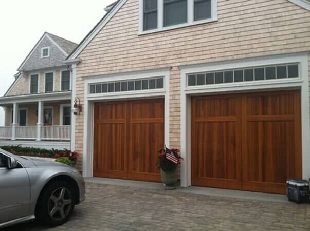 Two natural wood garage doors with top row windows on a shingled house exterior with a car parked in the driveway.