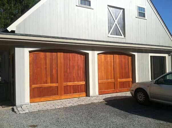 Two wooden arched garage doors with a light gray building exterior and a partial view of a car on the right.