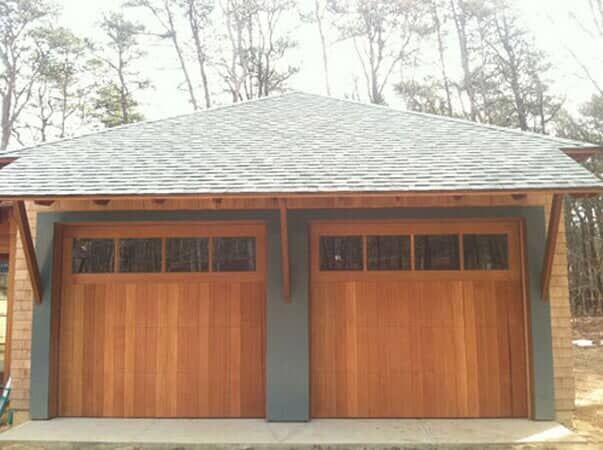 A two-car garage featuring wooden doors with windows, set beneath a gray shingled roof and wood-trimmed overhang.