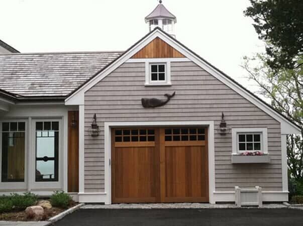 A light gray shingled garage with wooden doors, a whale wall ornament, a small window with a flower box, and a cupola.
