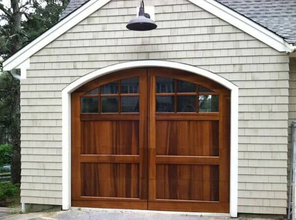 A two-car garage with a brown wooden, arched double door, set against light-colored shingled siding.