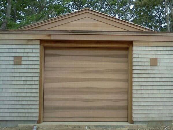 A modern wood-paneled garage door centered on a small structure with light-colored shake siding and a gabled roof.