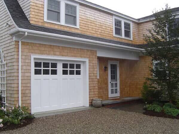 A house with light brown cedar shake siding, featuring a white garage door, a front door, and a gravel driveway.