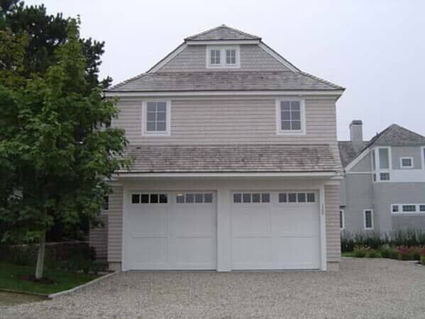 A light-gray, two-story carriage house with a cedar-shingle roof, two white garage doors, and a gravel driveway.