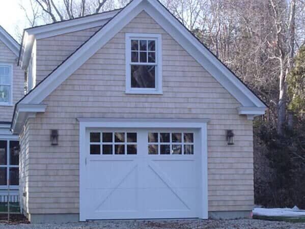A beige, shingled detached garage with a white, windowed carriage-style door and a single window in the gable.