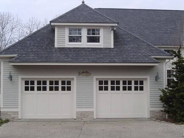 A two-car garage with white doors, light gray siding, a stone base, and a dormer window under a gray shingled roof.