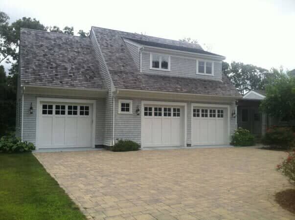 A light gray, three-car garage with white doors, shingles, and a paved driveway surrounded by trees.