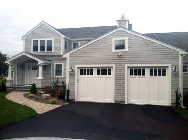 A two-story grey shingled house with a white front porch and an attached two-car garage with white paneled doors.