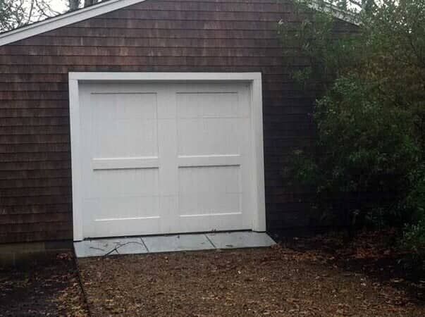 White double garage doors set in a brown shingled wall, with a small stone threshold leading to a dirt and leaf driveway.