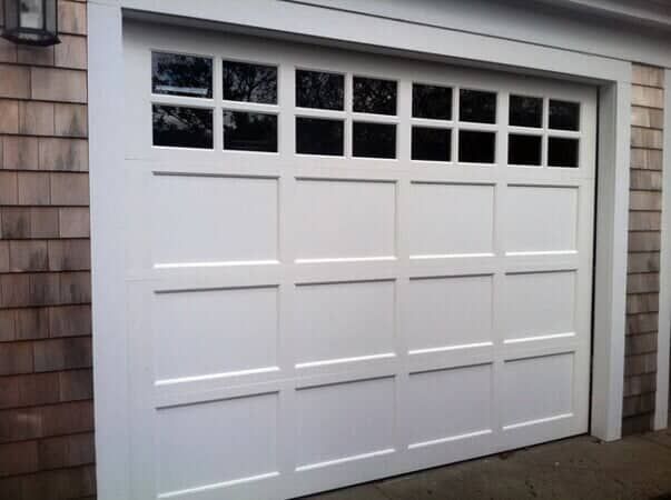 A white, four-panel carriage-style garage door with a row of small windows across the top, set against wood-shingled siding.