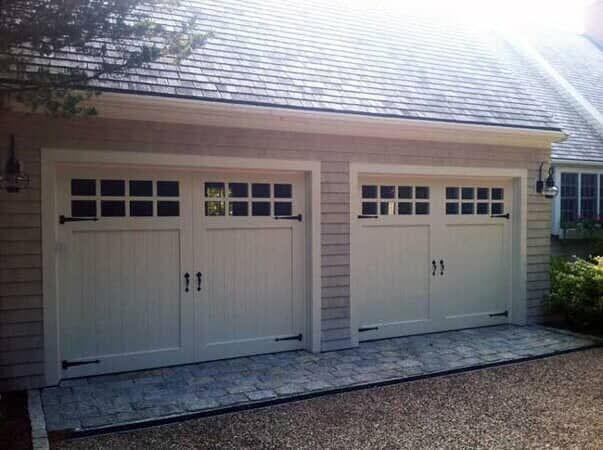 A side-by-side view of two cream-colored garage doors with windows and black hardware on a shingled building.