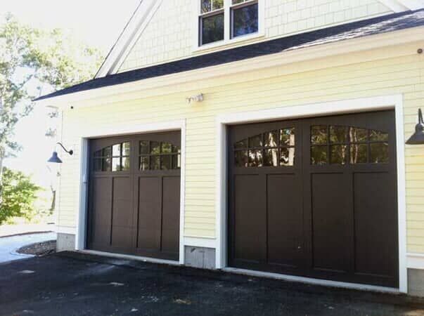 A yellow house with two dark brown garage doors featuring arched, multi-pane windows, set against a tree-lined driveway.