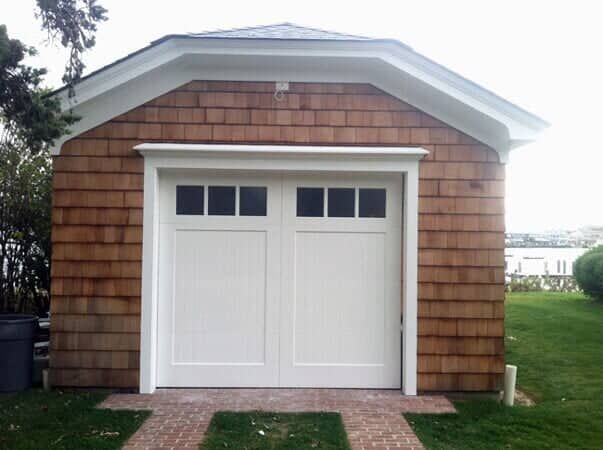 A white garage door with rectangular windows on a shingled building, with brick paths in front and a grassy lawn nearby.