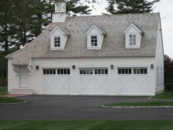 A white, three-bay garage with dormer windows and a small cupola sits on an asphalt driveway near trees.