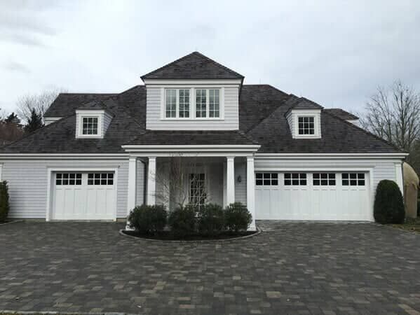 A grey house with a shingled roof, white trim, a centered portico, two garage doors, and a stone paved driveway.