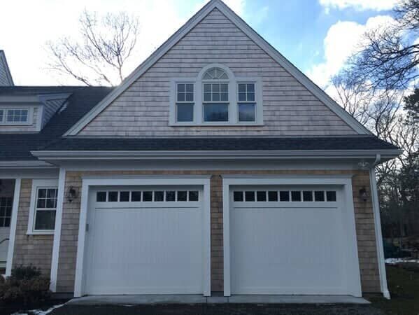 A two-car garage with white doors under a gabled roof with cedar shingles and a triple-pane attic window.
