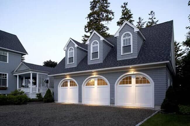 A gray, two-story house with a three-car garage, white arched doors, and three dormer windows on a gravel driveway.