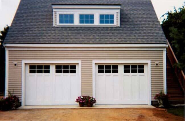 A detached two-car garage with white paneled doors, light tan siding, and a dormer window on the roof.