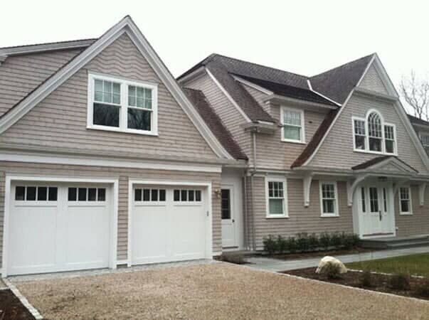 A two-story suburban house with light gray cedar shake siding, white trim, two garage doors, and a gravel driveway.