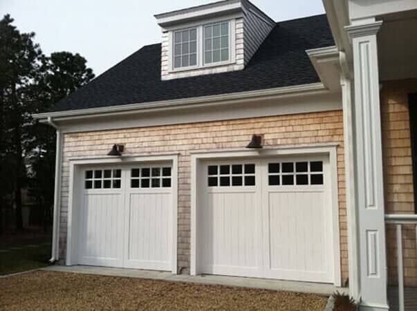 A two-car garage with white paneled doors, shingled siding, and a dormer window above on a residential home.
