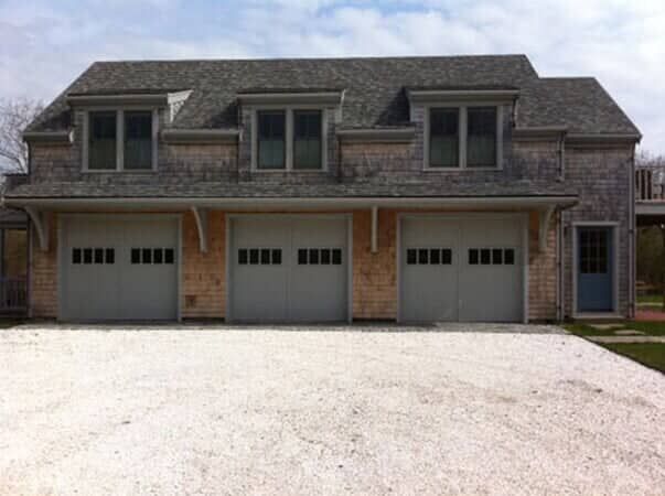 A three-car garage with shingled siding, a gray roof, three dormer windows, and a gravel driveway.