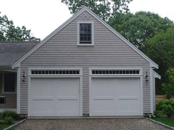 A detached, two-car garage with light-gray shingle siding, white doors with windows, and a single window in the gable.