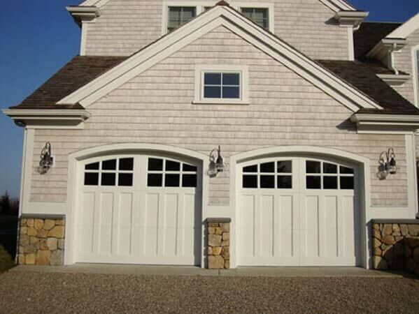 A two-car garage with white arched doors featuring window panes, beige shingle siding, and stone accents at the base.