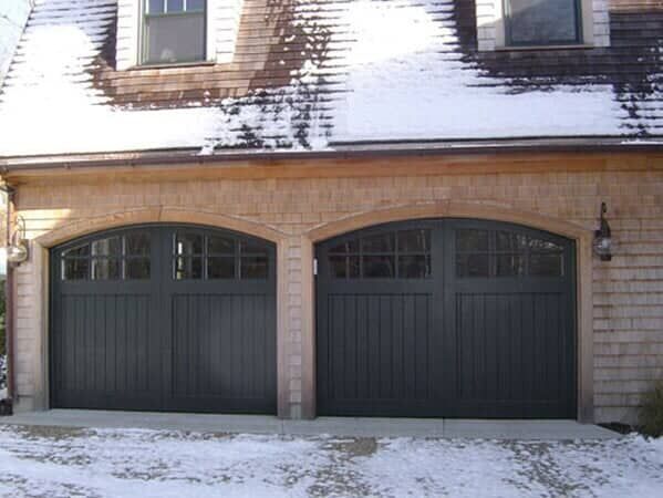 Two dark-colored arched garage doors with top windows, set in a stone-clad house exterior during a snowy winter.