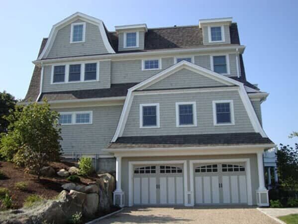 A light gray, shingled two-car garage house with a Gambrel roof on a sloped, rocky lot.