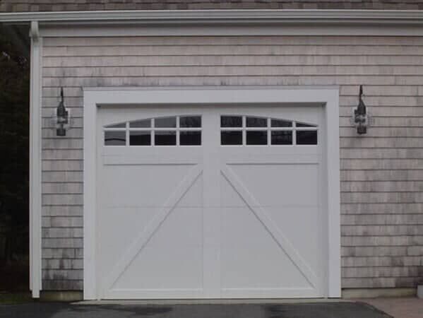 A white carriage-style garage door with arched windows and a diagonal brace design on a building with wood shingle siding.