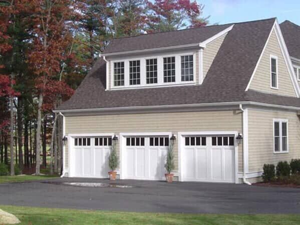 A tan, three-car garage with white doors, a gabled roof, and a large dormer window, set against a background of trees.