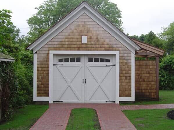 A detached garage with cedar shake siding, white carriage-style doors, and a brick driveway set in a green lawn.