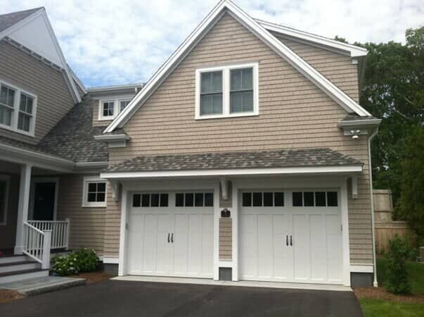 A two-car garage with white doors, light brown shingle siding, and a gabled roof, attached to a house with a front porch.