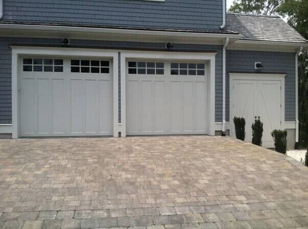 Gray house with two white carriage-style garage doors and a single side door, fronted by a stone paver driveway.