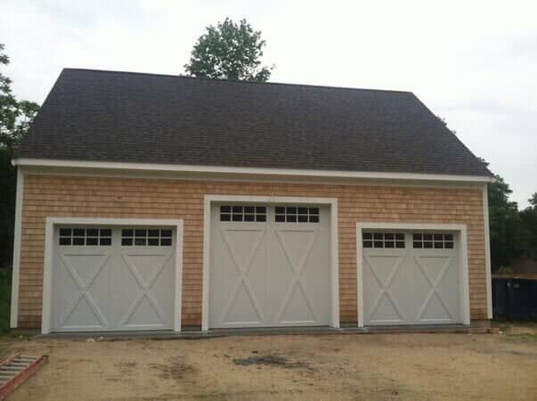 A detached garage with cedar shake siding, a dark roof, and three white carriage-style garage doors with window panes.