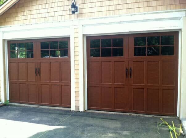 Two dark wood-toned, paneled garage doors with upper window grids and black handles set in a light-colored home exterior.