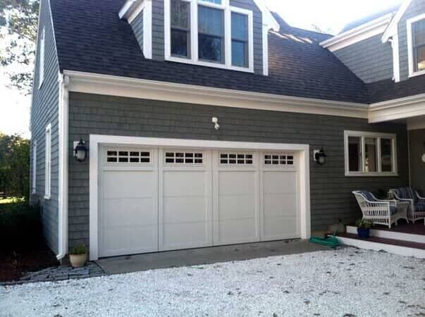 A grey shingled house with a large, four-section white garage door, a dormer window, and a white gravel driveway.