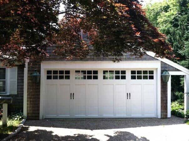 A white, carriage-style garage door with glass window panes sits in a brown-shingled garage under a large, leafy tree.
