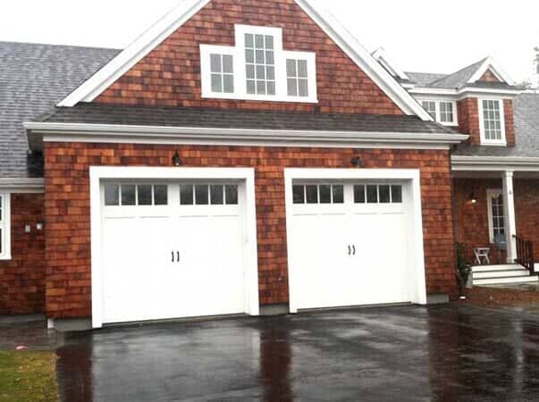 A two-car garage with white doors and windows, set in a house with wood-shingled siding on a rainy day.