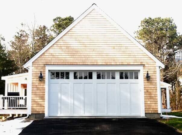 A garage with shingle siding, a white multi-pane door, and black lanterns on both sides, set against a wooded background.