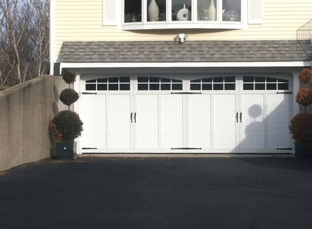 A white, carriage-style garage door with windows and decorative hardware, flanked by two topiary plants on a driveway.