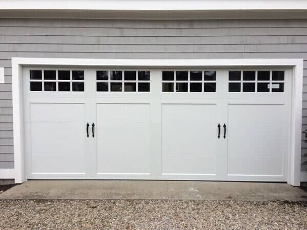 White carriage-style garage door with a four-section design, windows, and decorative black handles on a grey-sided home.