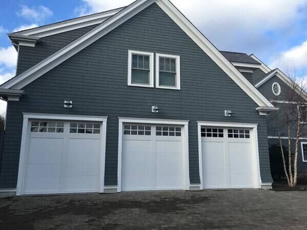 A blue-gray garage with three white carriage-style doors and two windows above under a bright, cloudy sky.
