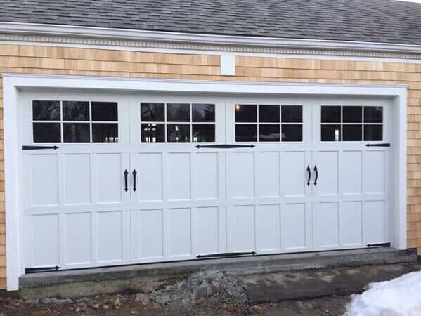 A white carriage-style garage door with windows across the top, decorative black hinges, and handles, set in a wood home.