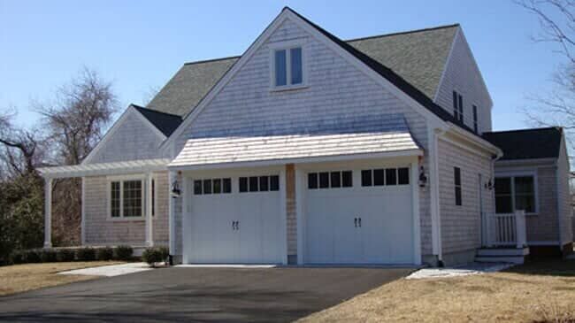 A two-story light gray shingled house with a two-car garage, white trim, and a small porch on a paved driveway.