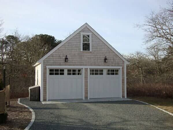 A detached two-car garage with light wood shingles, white trim, and a small single window, set on a gravel driveway.